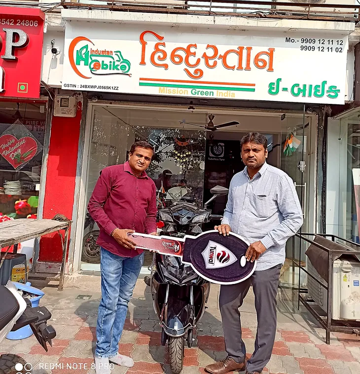 Two customers with a ceremonial key beside a newly delivered electric two-wheeler