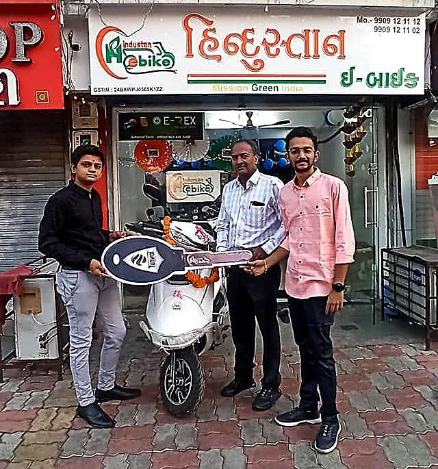 Customers collecting a new electric scooter outside the Hindustan E-Bike showroom in Sarthana, Surat, beneath the shop's Gujarati signboard
